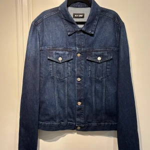 Jeansjacka BLK DNM - Mörkblå jeansjacka från BLK DNM med silverfärgade knappar, två bröstfickor. Jackan har en tidlös design med krage och långa ärmar, perfekt för dig som gillar en clean och stilren look.