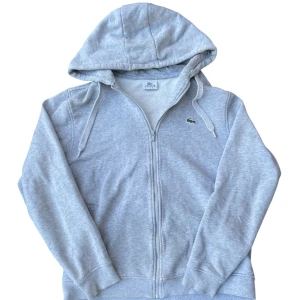 Grå hoodie från Lacoste med dragkedja - Snygg ljusgrå hoodie från Lacoste med klassisk krokodillogga på bröstet. Tröjan har huva med snörning, hel dragkedja och ribbade muddar. Perfekt för en chill och sportig stil. Tillverkad i mjuk bomullsmix för extra komfort. Har hål fram på armarna men inte så märkbara.