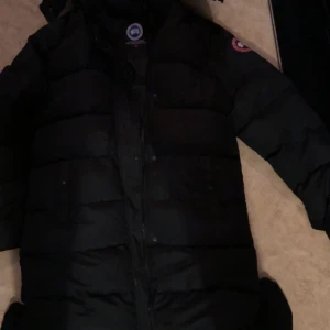 Svart lång dunjacka från Canada Goose - Säljer en svart dunjacka från Canada Goose med klassisk patch på ärmen. Jackan är lång, har huva och dragkedja med tryckknappar framtill. Perfekt för kalla vinterdagar och har en clean, stilren look som funkar till allt.