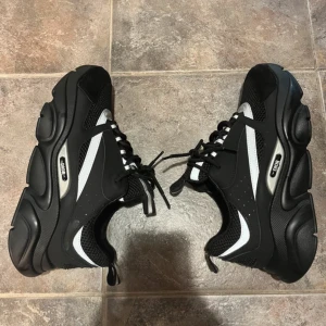 Svarta Dior B22 sneakers med reflex - Säljer ett par chunky Dior B22 sneakers i svart med silvriga och vita reflexdetaljer. Skorna har mesh och skinnpaneler, kraftig platt sula och snörning. Snygg sportig vibe med exklusiv känsla och tydlig Dior-logga på sidan av sulan.