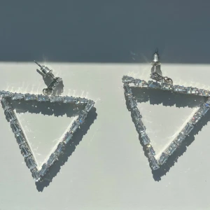 Glittriga trekantsörhängen i silver - Snygga örhängen i form av stora trianglar med glittrande stenar runt hela kanten. De är silvriga och har en modern, edgy vibe som verkligen drar blickarna till sig. Perfekta för dig som vill sticka ut med accessoarerna.