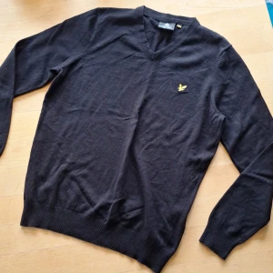 Svart v-ringad tröja från Lyle & Scott - Svart v-ringad tröja från Lyle & Scott med klassisk gul broderad logga på bröstet. Tröjan är stickad och har långa ärmar samt ribbade muddar vid ärmslut och nederkant. Perfekt för lager-på-lager och snygg till jeans.