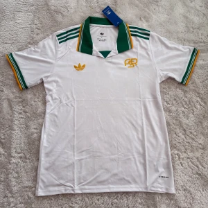 Vit Adidas fotbollströja med gröna detaljer - Snygg vit fotbollströja från Adidas med grönt och gult på krage och ärmslut. Klassisk retrostil med broderad Adidas-logga och AR-emblem i gult på bröstet. Tillverkad i lätt, ventilerande polyester med diskret mönster i tyget. Perfekt för dig som gillar sportig stil.