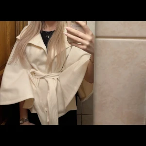 Poncho - Säljer en poncho med bred krage och knytbälte i midjan. Jackan har en snygg omlottstil och vida ärmar som ger en cool och elegant look. Perfekt för dig som gillar stilrena och trendiga plagg.