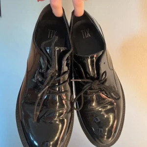 Svarta lackade chunky loafers XIT - Säljer ett par svarta loafers från TIX med glansig lackad yta och kraftig, räfflad sula. Skorna har snörning framtill och en klassisk rund tå. Perfekta för dig som gillar en edgy och stilren look.