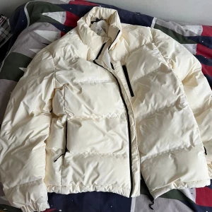 Vit pufferjacka med svarta detaljer - Säljer en stilren vit pufferjacka från H&M med svarta dragkedjor och hög krage. Jackan är vadderad och har två sidofickor samt en bröstficka med dragkedja. Perfekt för kalla dagar när du vill hålla värmen och ändå se fräsch ut.