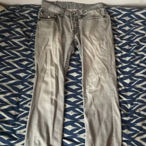 Grå True Religion Billy SuperT jeans - Säljer ett par grå jeans från True Religion, modell Billy SuperT. Jeansen har raka ben, klassiska kontrastsömmar och stora bakfickor med lock och knapp. Tillverkade i USA och i bomullsmaterial. Perfekta för dig som gillar streetwear och vill sticka ut. De har ett hål i framfickan åt höger men annars helt okej skick, mycket kvar att ge