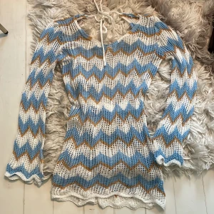 Stickad zigzagklänning i blått och vitt - Superfin stickad strand kortklänning med zigzag-mönster i blått, vitt och brunt. Klänningen har lång ärm, v-ringning med knyt och är virkad i ett luftigt material. Perfekt för sommarens alla tillfällen och ger en riktigt trendig vibe.