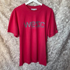 Röd t-shirt från WESC i bomull - Röd t-shirt från WESC med grå logga framtill. Klassisk rund halsringning och korta ärmar. Tillverkad i mjuk bomull och har en liten patch på ärmen. Tyget är lite urtvättat.