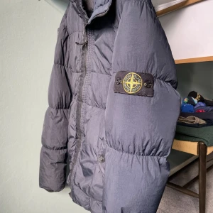 Mörkgrå pufferjacka från Stone Island - Säljer en mörkgrå pufferjacka från Stone Island med ikonisk patch på ärmen. Jackan har huva, dragkedja framtill och två fickor med knappstängning. Perfekt för kalla dagar och har en clean, urban vibe.  Det är i storlek S passade mig perfekt när ja va 175 så skulle säga passar de som e mellan 170-180 cm lång. Är i fint skick använd förra vintern. Säljs då för liten. Enbart ett litet hål på högra axeln. Köpt för runt 1500. QR kod finns att scanna ( se bild 5). Priset ej hugget i sten.