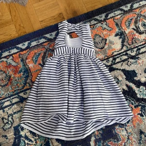 Randig sommarklänning med rosett - Superfin kortklänning med marinblå och vita ränder, söt rosett i nacken och öppen rygg. Klänningen har knappar i halsen och ett fluffigt vitt foder som ger extra volym. Perfekt för varma dagar och har en klassisk, fräsch look.