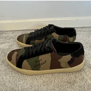 Saint Laurent sneakers i camouflage - Snygga sneakers från Saint Laurent med klassiskt camouflage-mönster i grönt, brunt och beige. Skorna har svarta snören, svarta detaljer och en beige sula. Saint Laurent-loggan är tryckt på sidan. Perfekta för dig som vill sticka ut med en cool streetstyle.