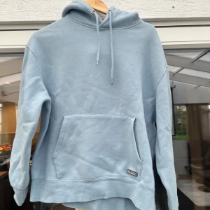 Ljusblå hoodie från Pull&Bear - Säljer en ljusblå hoodie från Pull&Bear med klassisk känguruficka och snörning i huvan. Mjuk och skön, perfekt för chill dagar. Enkel design utan mönster och med en liten logga på fickan.