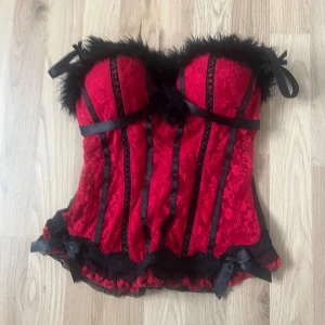 Röd och svart korsett med spets och fjädrar - Snygg röd korsett med svarta detaljer, spets och fjäderkant upptill. Dekorativa svarta band och rosetter, samt genomskinliga partier baktill. Säljer för att jag inte gillar hur den sitter och den kommer ej till användning 