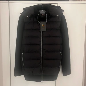 Svart pufferjacka från Leon Marcel - Stilren svart pufferjacka från Leon Marcel med stickade ärmar och vadderad kropp. Jackan har avtagbar luva med dragkedja. Perfekt för kyliga dagar. Längd: 67cm
