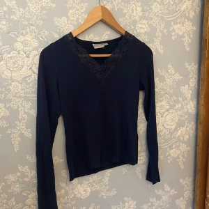 Mörkblå långärmad topp med spets - Mörkblå långärmad topp från Gina Tricot med snygg v-ringning dekorerad med spets. Toppens material är mjukt och stretchigt, perfekt för en stilren look. Passformen är normal och toppen är enkel att matcha med både jeans och kjol.