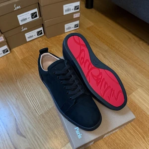 Svarta mocka sneakers från Louboutin - Säljer ett par svarta sneakers från Christian Louboutin i mjuk mocka med klassisk röd sula och rund tå. Skorna har svarta snören och diskreta detaljer, samt en platt sula. Insidan är ljusbeige och designen är stilren och lyxig med Paris-vibe.
