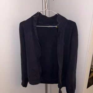 Mörkblå kofta från Polo Ralph Lauren - Mörkblå stickad kofta från Polo Ralph Lauren med dragkedja framtill och diskret broderad logga på bröstet. Koftan har ribbade partier vid axlar och krage samt två fickor framtill. Perfekt för lager-på-lager under kyliga dagar.