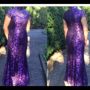 Lång lila spetsklänning med hög krage - Elegant långklänning i lila spets med hög krage och korta ärmar. Klänningen har ett vackert blommigt spetsmönster och figurnära passform som framhäver siluetten. Perfekt för speciella tillfällen när du vill sticka ut.