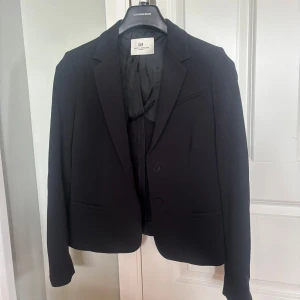 Svart kavaj / Blazer  från DAY Birger et Mikkelsen - Stilren svart kavaj från DAY Birger et Mikkelsen med klassisk krage, två knappar framtill och diskreta fickor. Kavajen har en figurnära passform och är helfodrad för en snygg siluett. Perfekt för dig som gillar en clean och tidlös look.