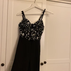 Svart byxdress med paljetter - Snygg svart byxdress med smala axelband och vida ben. Överdelen är täckt av glittriga paljetter i silver som ger en riktigt festlig look. Perfekt för dig som vill sticka ut och glänsa. Materialet känns mjukt och följsamt. Runt bröstet har själva taggen kommit ut men man kan nog sy ihop den igen, säljer för att varan är för liten annars är den jätte fin 