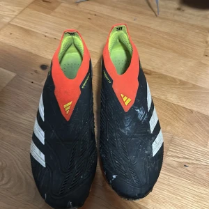 Adidas Predator fotbollsskor svart/röd - Säljer ett par Adidas Predator fotbollsskor i svart med vita detaljer och röda samt gula inslag. Skorna har en strumpliknande passform utan snörning, mönstrad ovandel och dobbar för gräsplan. Snygg och modern design, perfekt för dig som vill sticka ut på planen.
