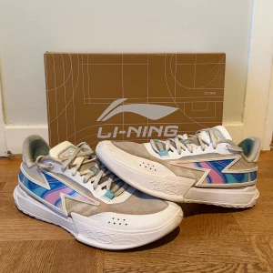 Li-Ning Way Of Wade Son Of Flash - Säljer ett par Li-Ning Way Of Wade Flash sneakers i vitt med inslag av blått, rosa och silverdetaljer. Skorna har en chunky sula, snörning och coolt mönstrad sidopanel. Materialmix av textil och TPU ger en sportig vibe. Perfekta för dig som gillar unika och färgglada sneakers. 