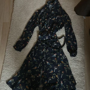 Mörkblå långklänning med fågelmönster Bondelid - Supersnygg långklänning från Bondelid i mörkblått med färgglatt fågel- och bladmotiv. Klänningen har långa ärmar, knytband i midjan och en flowy passform. Perfekt för dig som gillar unika prints och vill sticka ut lite extra. 