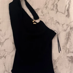 Svart ribbad oneshoulder topp från Zara - Säljer en svart ribbad oneshoulder topp från Zara med snygga guldfärgade metalldetaljer på axelbandet. Toppen har en tight passform och är perfekt för dig som vill sticka ut med en cool och modern look.