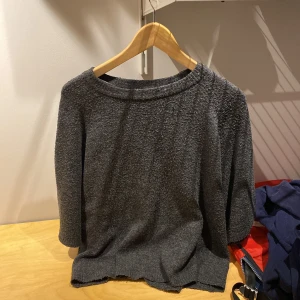 Mörkgrå stickad tröja från 157 - Mysig mörkgrå stickad tröja från 157 med trekvartsärm och rund halsringning. Tröjan har en enkel och avslappnad passform, perfekt för lager på lager. Materialet är mjukt och ger en chill vibe till din outfit.