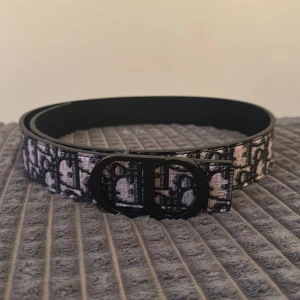 Svart och beige bälte - Snyggt bälte  med klassiskt monogrammönster i svart och beige. Bältet har en stilren, svart spänne med rundade former och är tillverkat i skinn med textil på ovansidan. Perfekt accessoar för att lyfta din outfit.