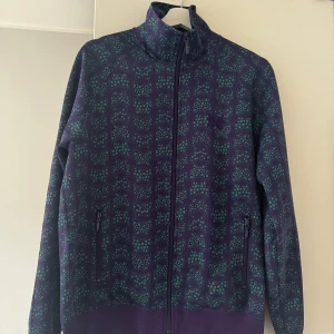 Lila mönstrad jacka från Needles M - Säljer en unik jacka från Needles i storlek M. Jackan är lila med grönt fjärilsmönster, har hög krage, dragkedja framtill och två fickor med dragkedja. Materialet är troligtvis polyester och passformen är normal. Perfekt för dig som vill sticka ut med coola detaljer.den är köpt på caliroots. Mer eller mindre i nyskick!