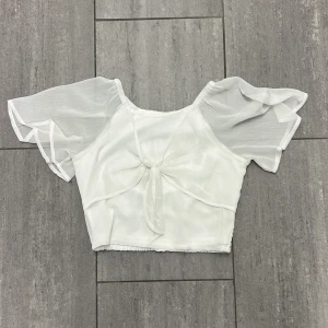 Vit blus med knyt från Lindex - Söt vit blus från Lindex med kort, croppad passform och luftiga, transparenta ärmar. Toppen har en snygg knytning framtill och smockad nederkant bak. Perfekt för en fräsch och somrig look.
