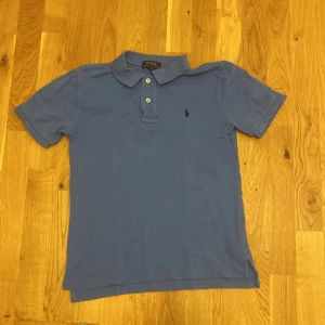 Blå pikétröja från Ralph Lauren - Klassisk blå pikétröja från Polo Ralph Lauren med korta ärmar och krage. Tröjan har knäppning med två knappar och den ikoniska broderade loggan på bröstet. Perfekt för en stilren och avslappnad look. Vid snabb affär kan priset ändras 