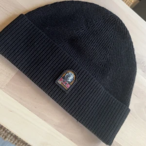 Svart ribbstickad mössa Parajumpers - Svart ribbstickad mössa från Parajumpers med klassisk uppvikt kant och broderad PJS-logga framtill. Mössan är tillverkad i mjukt material och har en röd etikett med Parajumpers på insidan. Perfekt för kyliga dagar och snygg till streetwear.