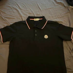 Svart Moncler pikétröja med logga - Svart pikétröja från Moncler med klassisk krage och tre knappar. Tröjan har Moncler-logga på bröstet och snygga rödvita ränder på krage och ärmslut. Perfekt för dig som vill ha en stilren och sportig look.