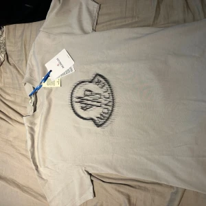  Moncler t-shirt med logga - Snygg beige t-shirt från Moncler med stort svart logotryck på bröstet och en liten svart patch på ärmen. Klassisk rund hals och korta ärmar. Tillverkad i mjuk bomull för en skön känsla. Perfekt för dig som gillar stilrena plagg med exklusiv känsla. HELT NY OCH ÄKTA!!