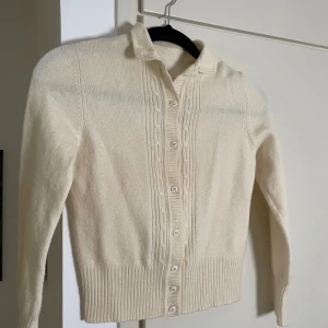 Creméfärgad ullkofta med knappar - Creméfärgad stickad ullkofta med kabelstickat mönster längs knappraden. Sparsamt använd och i fint skick. Storlek XS. 