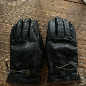 Svarta skinnhandskar från Hestra - Snygga svarta handskar från Hestra i getskinn med bruna detaljer vid mudden och justerbar dragsko. Insidan är fodrad med mjuk polyester för extra komfort. Perfekta för kalla dagar när du vill hålla stilen och värmen.