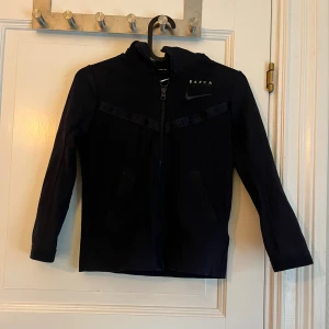 Svart Nike Barca hoodiejacka XS - Svart hoodiejacka från Nike med Barca-tryck på bröstet. Jackan har dragkedja framtill, huva och två fickor. Snygga detaljer med svarta band och tryckknapp bak i nacken. Perfekt för dig som gillar sportig stil.