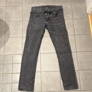 Grå jeans från Nudie Jeans Co - Snygga grå jeans från Nudie Jeans Co med klassisk femficksdesign och svarta detaljer på bakfickorna. Jeansen har raka ben och knäppning med knappar framtill. Perfekt för dig som gillar en stilren och avslappnad look.