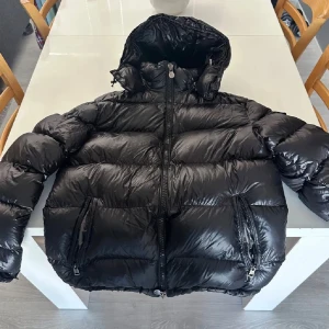 Svart Moncler pufferjacka med huva - Säljer en svart pufferjacka från Moncler med glansig finish och stor huva. Jackan har dragkedja framtill, två fickor med dragkedja och en extra ficka med Moncler-logga på ärmen. Perfekt för kalla dagar och riktigt snygg streetstil. Den passar till både M-L. Den har skador och det är därför jag säljer den så billig. NFC och allt annat fungerar också.