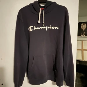 Svart hoodie från Champion, stl L - Säljer en svart hoodie från Champion i storlek L. Tröjan har vit logga framtill, huva med vita snören och en stor magficka. Perfekt för dig som gillar en klassisk streetwear-look. Materialet är mjukt och skönt, passar till chill dagar.