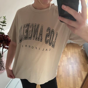 Beige oversized t-shirt Los Angeles - Säljer en beige oversized t-shirt med svart tryck 'Los Angeles California' på bröstet. T-shirten har rund halsringning och korta ärmar. Perfekt för en chill och avslappnad stil. Materialet känns mjukt och skönt mot huden.