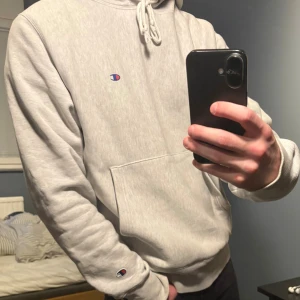 Ljusgrå hoodie från Champion - Säljer en stilren ljusgrå hoodie från Champion med klassisk logga på bröstet och ärmslutet. Tröjan har huva med snörning och en stor magficka. Perfekt för dig som gillar en enkel och clean look.