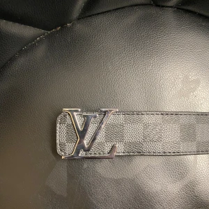 Louis Vuitton Damier Graphite bälte - Snyggt Louis Vuitton bälte i Damier Graphite-mönster med klassiskt LV-spänne i silver. Bältet är svart och grått med rutigt mönster och tillverkat i skinn. Perfekt accessoar för att lyfta din outfit med en lyxig touch.