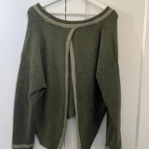 Olivgrön stickad tröja med öppen rygg - Olivgrön stickad tröja med ljusare kantdetaljer och snörning framtill. Tröjan har en ovalformad passform och en öppen fin rygg!