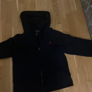Svart hoodie från Polo Ralph Lauren - Svart hoodie från Polo Ralph Lauren med dragkedja och klassisk röd logga broderad på bröstet. Tröjan har huva, långa ärmar och är tillverkad i mjuk bomull. Perfekt för en chill och stilren look.