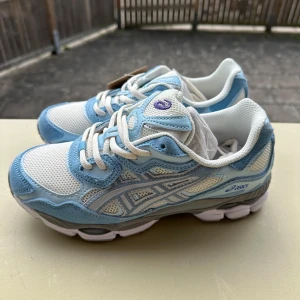 Asics GEL-NYC i blått och vitt - Snygga Asics GEL sneakers i vitt och ljusblått med mesh och mockadetaljer. Skorna har klassisk snörning, rund tå och en chunky sula som ger en sportig vibe. Perfekta för dig som gillar streetstyle och bekväma skor med cool design.