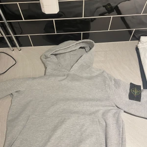 Grå hoodie från Stone Island - Säljer en stilren grå hoodie från Stone Island med klassisk logga på ärmen. Tröjan har huva, känguruficka och är tillverkad i mjuk bomull. Perfekt för dig som gillar streetwear och vill ha något bekvämt och snyggt.